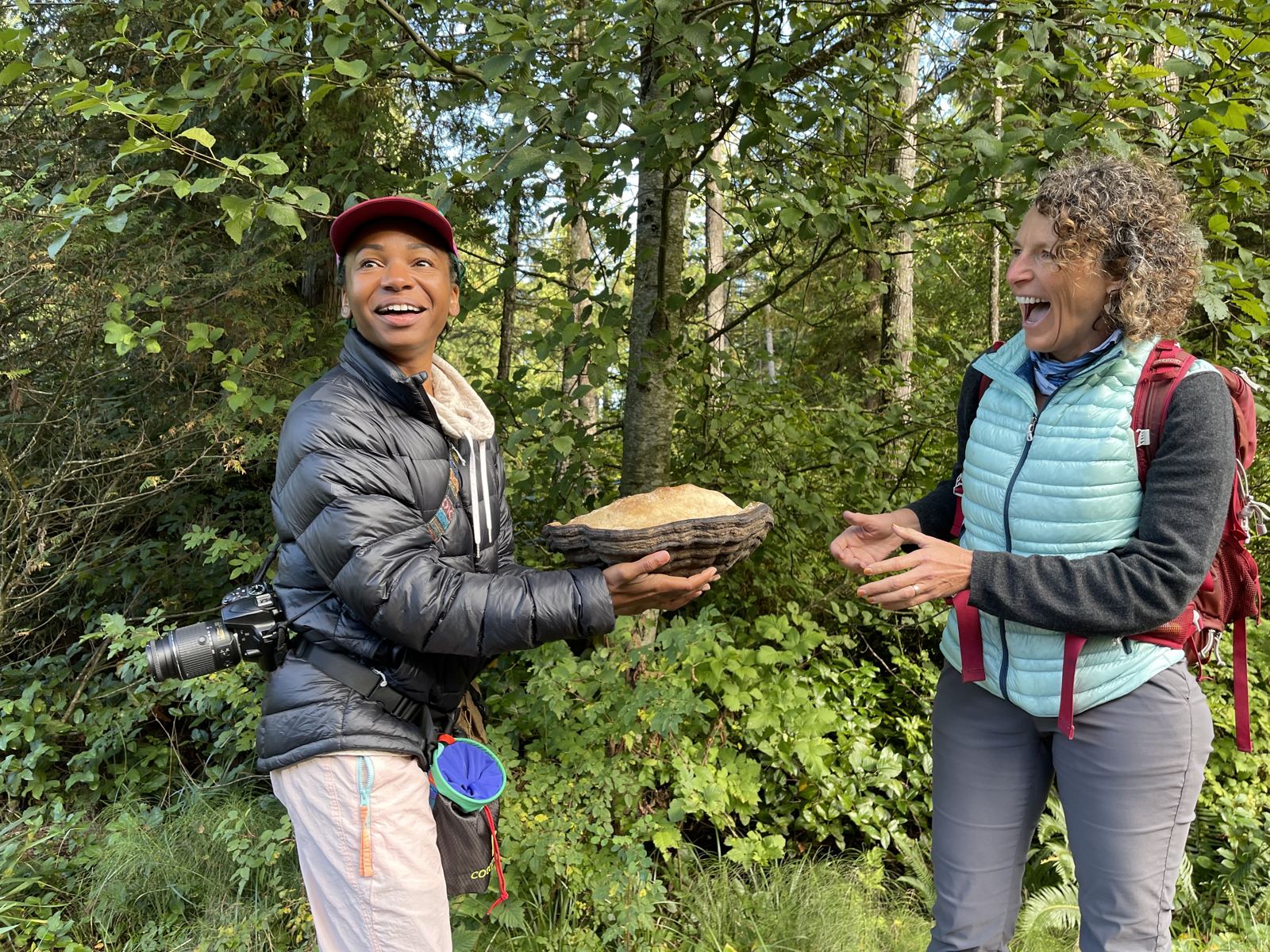 Two naturalists in a forest laughing as one holds out a large bracket fungus.